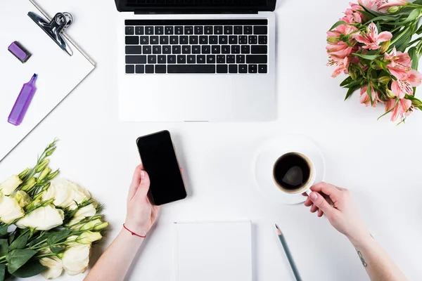 Vista recortada de la mujer sosteniendo teléfono inteligente con pantalla en blanco y taza de café en el lugar de trabajo - foto de stock