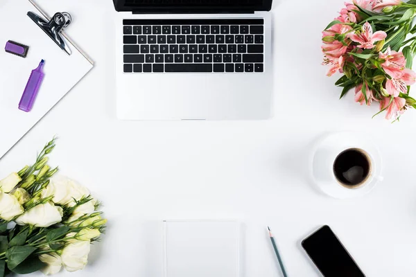 Draufsicht auf Laptop, Smartphone mit leerem Bildschirm, Kaffeetasse, Klemmbrett mit Textmarker, Buch, Bleistift und Blumen auf Weiß — Stockfoto