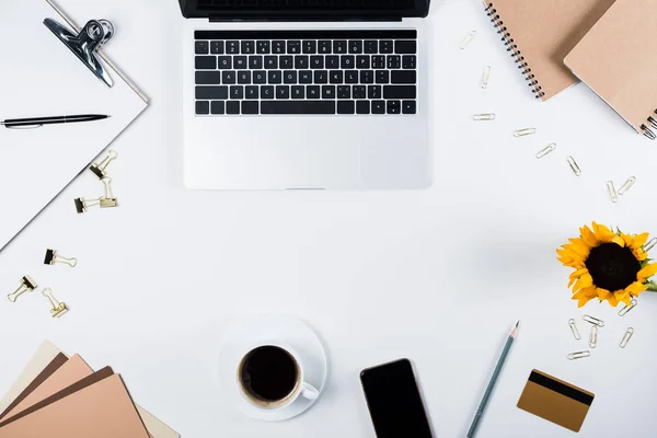 Top view of laptop, smartphone with blank screen, sunflower, clipboard, cup of coffee, craft paper, credit card, binder clips and paper clips on white — Stock Photo