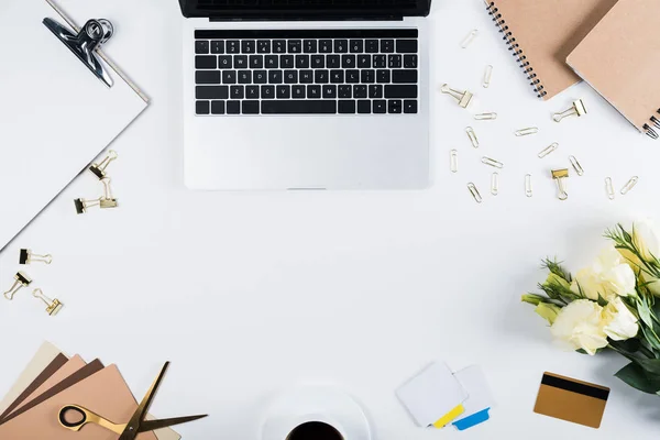 Top view of laptop, clipboard, cup of coffee, notebooks, craft paper, credit card, flowers, scissors, binder clips and paper clips on white — Stock Photo