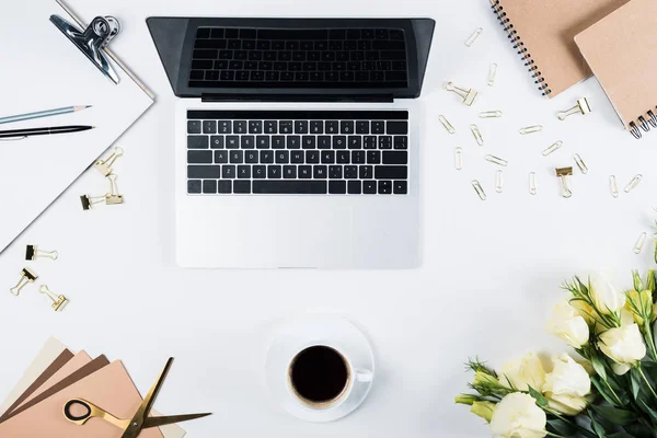 Vista dall'alto del computer portatile con schermo bianco, tazza di caffè, appunti, fiori, penna, matita, forbici, clip legante e graffette di bianco — Foto stock