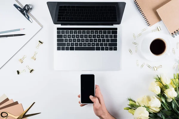 Vista ritagliata dell'uomo che tiene smartphone con schermo vuoto vicino al computer portatile sul posto di lavoro — Foto stock