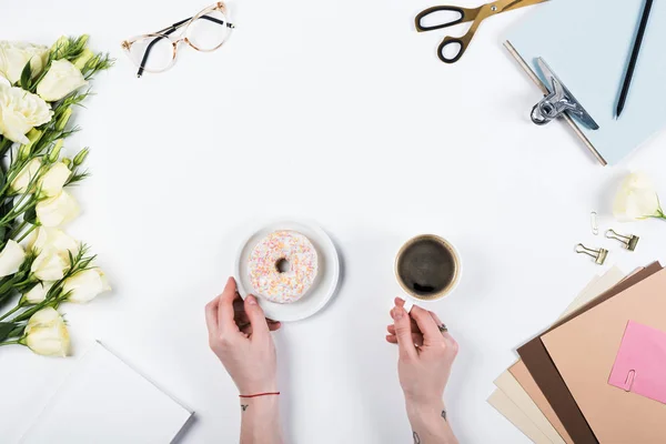 Cropped view of woman with doughnut and cup of coffee at workplace — Stock Photo
