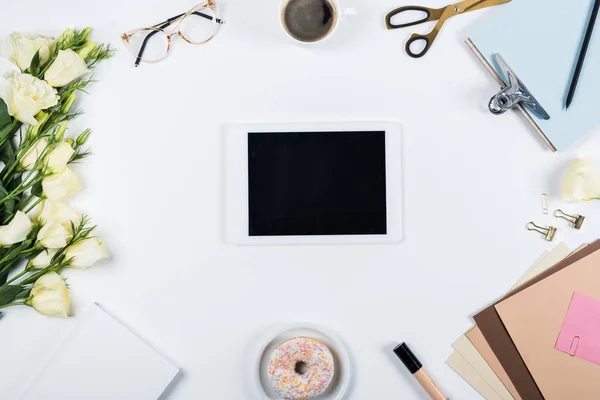 Vista dall'alto del tablet digitale con schermo bianco, tazza di caffè, ciambella, fiori, occhiali, forbici, carta artigianale, appunti e correttore su bianco — Foto stock