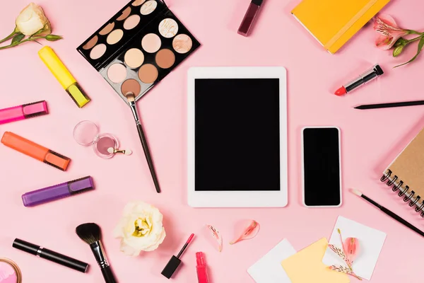 Вид зверху цифрового планшета та смартфона з порожнім екраном, блокнотами, квітами та декоративною косметикою на рожевому — стокове фото