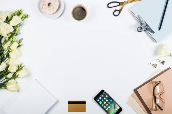 KYIV, UKRAINE - MAY 11, 2019: top view of cup of coffee, doughnut, credit card, flowers, glasses, scissors, clipboard and smartphone with home page on screen on white — Stock Photo