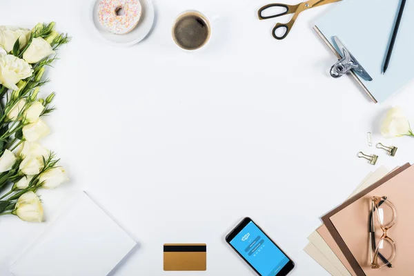 KYIV, UKRAINE - MAY 11, 2019: top view of cup of coffee, doughnut, credit card, flowers, glasses, scissors, clipboard and smartphone with skype app on screen on white — Stock Photo