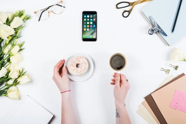 KYIV, UKRAINE - 11 MAI 2019 : vue recadrée de la femme avec tasse de café, beignet et smartphone avec page d'accueil à l'écran sur le lieu de travail — Photo de stock