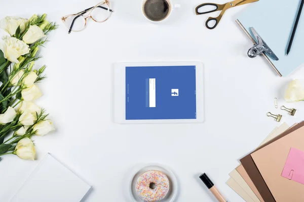 KYIV, UKRAINE - MAY 11, 2019: top view of cup of coffee, doughnut, flowers, glasses, book, papers, clipboard and digital tablet with facebook app on screen on white — Stock Photo