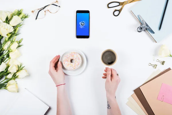 KYIV, UKRAINE - MAY 11, 2019: cropped view of woman with cup of coffee, doughnut and smartphone with shazam app on screen at workplace — Stock Photo