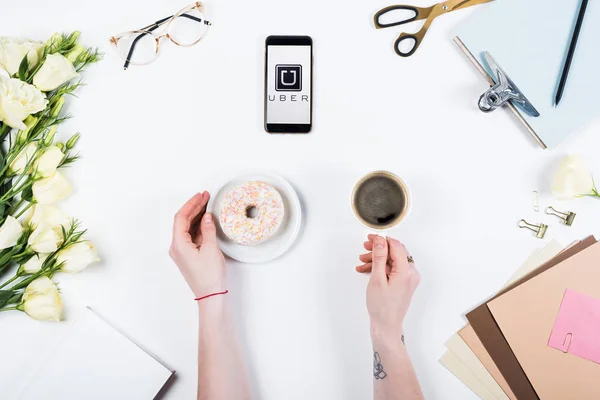 KYIV, UCRANIA - 11 de mayo de 2019: vista recortada de la mujer con taza de café, donut y teléfono inteligente con la aplicación uber en la pantalla en el lugar de trabajo - foto de stock