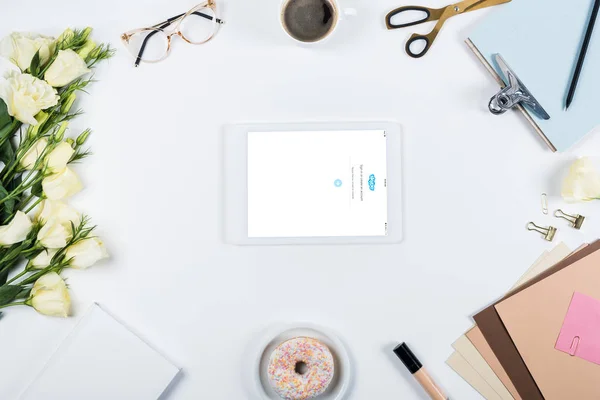 KYIV, UKRAINE - MAY 11, 2019: top view of cup of coffee, doughnut, flowers, glasses, book, papers, clipboard and digital tablet with skype app on screen on white — Stock Photo