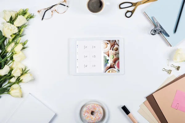 KYIV, UKRAINE - 11 MAI 2019 : vue du dessus de la tasse de café, beignet, fleurs, verres, livre, papiers, presse-papiers et tablette numérique avec application quadruple à l'écran sur blanc — Photo de stock