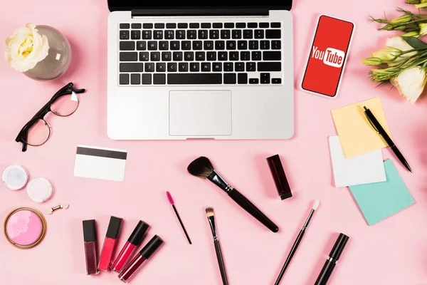 KYIV, UKRAINE - MAY 11, 2019: top view of laptop, smartphone with youtube app on screen, flowers, credit card and decorative cosmetics on pink — Stock Photo