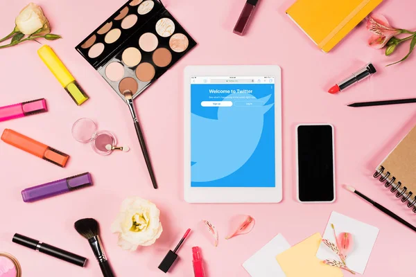 KYIV, UCRANIA - 11 de mayo de 2019: vista superior de la tableta digital con la aplicación de twitter en la pantalla, teléfono inteligente con pantalla en blanco, flores, iluminadores y cosméticos decorativos en rosa - foto de stock