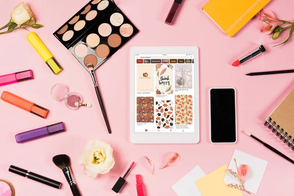 KYIV, UKRAINE - MAY 11, 2019: top view of digital tablet with pinterest app on screen, smartphone with blank screen, flowers, highlighters and decorative cosmetics on pink — Stock Photo