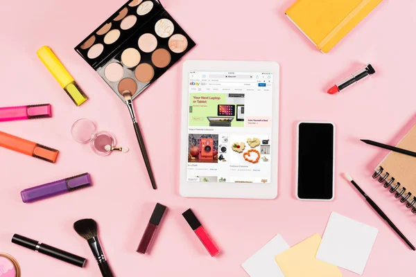 Вид зверху цифровий планшет з додатком eBay на екрані, смартфон з порожнім екраном, запальнички та декоративна косметика на рожевому — стокове фото