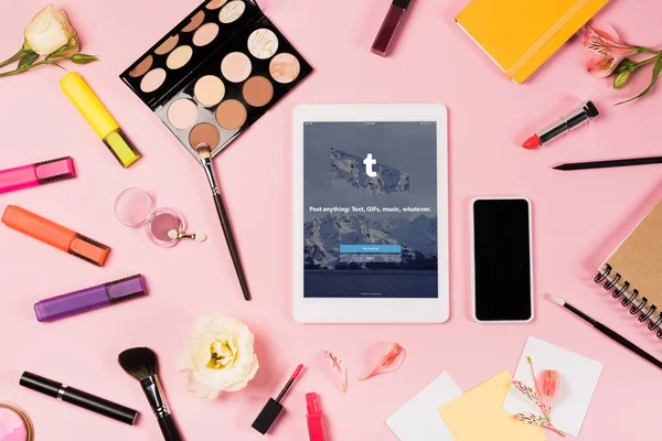 KYIV, UCRANIA - 11 de mayo de 2019: vista superior de la tableta digital con aplicación tumblr en la pantalla, smartphone con pantalla en blanco, flores, iluminadores y cosméticos decorativos en rosa - foto de stock