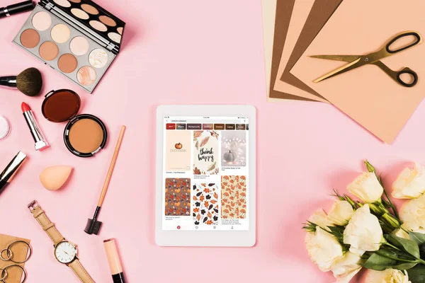 KYIV, UKRAINE - MAY 11, 2019: top view of digital tablet with pinterest app on screen, decorative cosmetics, flowers, craft paper and scissors on pink — Stock Photo