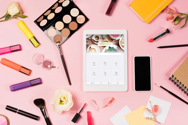 KYIV, UCRANIA - 11 de mayo de 2019: vista superior de la tableta digital con aplicación cuadrada en la pantalla, teléfono inteligente con pantalla en blanco, flores, iluminadores y cosméticos decorativos en rosa - foto de stock