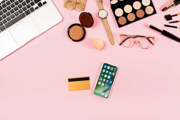 KYIV, UKRAINE - MAY 11, 2019: top view of laptop, decorative cosmetics, glasses, wristwatch, credit card and smartphone with lock screen on pink — Stock Photo