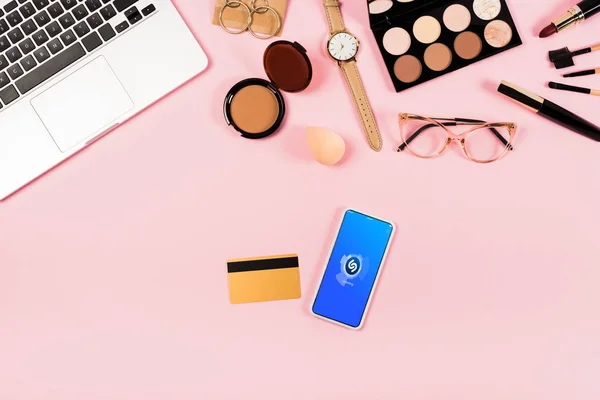 KYIV, UKRAINE - MAY 11, 2019: top view of laptop, decorative cosmetics, glasses, wristwatch, credit card and smartphone with shazam app on screen on pink — Stock Photo