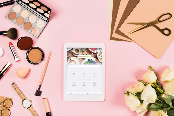 KYIV, UKRAINE - MAY 11, 2019: top view of decorative cosmetics, eustoma bouquet, craft paper with scissors and digital tablet with foursquare app on screen on pink — Stock Photo