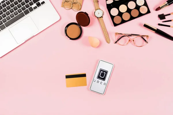 KYIV, UKRAINE - MAY 11, 2019: top view of laptop, decorative cosmetics, glasses, wristwatch, credit card and smartphone with uber app on screen on pink — Stock Photo
