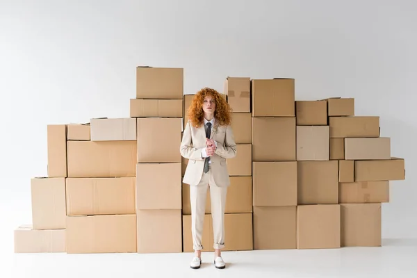 Attractive curly redhead girl looking at camera while standing near boxes on white — Stock Photo