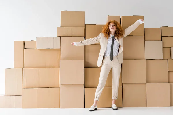 Atractiva pelirroja rizada de pie con las manos extendidas cerca de cajas en blanco - foto de stock