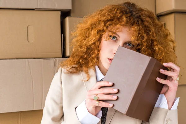 Redhead woman covering face with book near cardboard boxes — Stock Photo