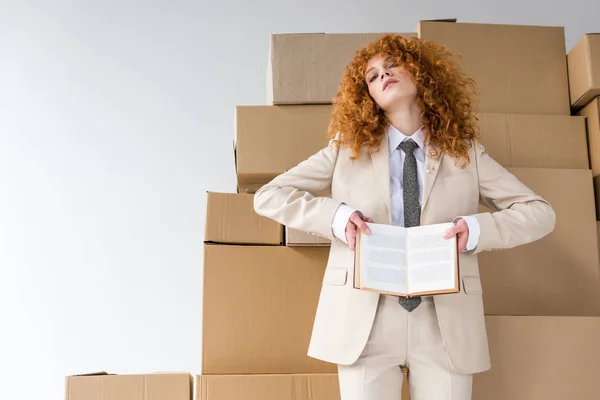 Low angle view of attractive redhead girl holding book near boxes on white — Stock Photo