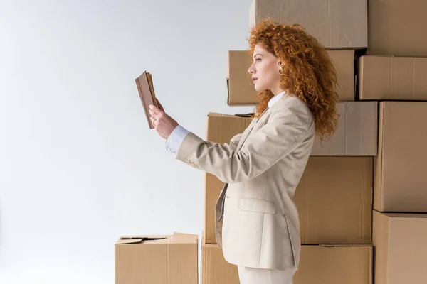 Attractive redhead girl reading book near brown boxes on white — Stock Photo
