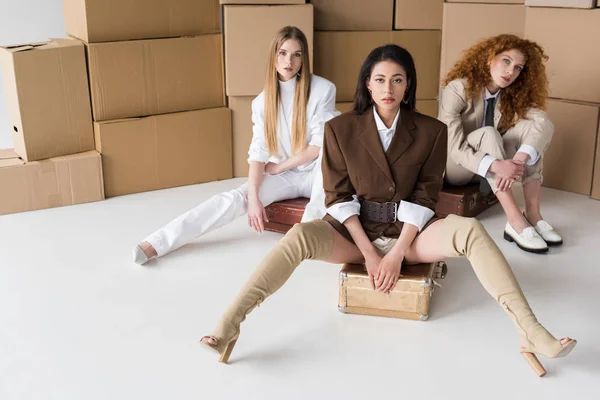 Belles filles multiculturelles assis sur des valises près de boîtes sur blanc — Photo de stock