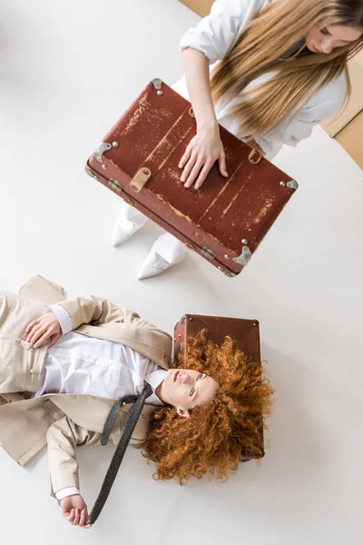 Вид зверху на руду жінку, що лежить поруч з блондинкою, що стоїть з валізою на білому — стокове фото