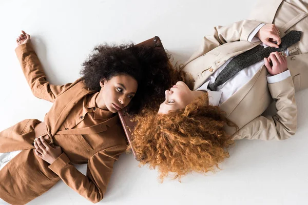 Top view of redhead girl lying near african american woman and suitcase on white — Stock Photo