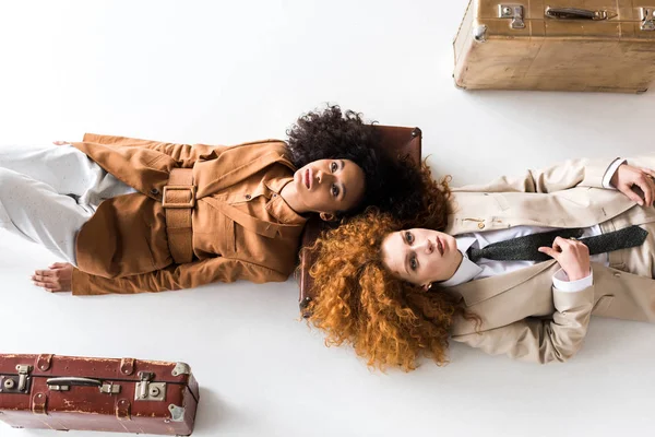 Top view of redhead girl lying near african american curly woman and suitcases on white — Stock Photo