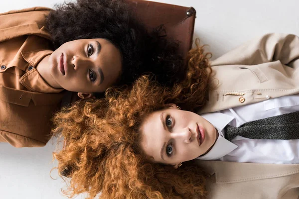 Vista superior de chica pelirroja acostada con mujer rizada afroamericana en bolsa de viaje en blanco - foto de stock