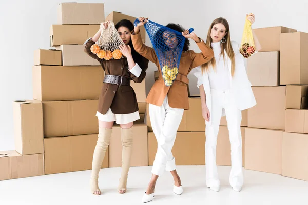 Afro-américaine filles couvrant les visages tout en tenant des sacs à ficelle avec des fruits près femme blonde sur blanc — Photo de stock