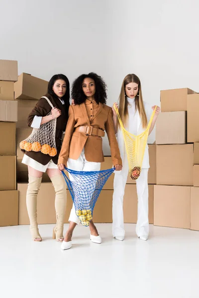 African american girls holding string bags with fruits near blonde woman and boxes on white — Stock Photo
