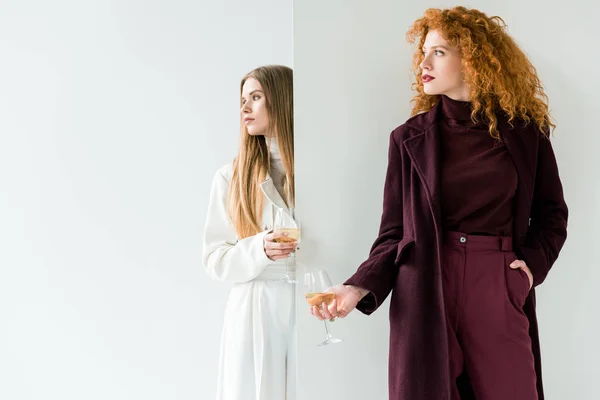 Attractive curly girl standing with hand in pocket and holding champagne glass near blonde woman on white — Stock Photo