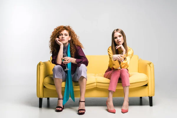 Redhead girl sitting on sofa near blonde woman with champagne glasses on white — Stock Photo