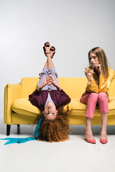 Rousse fille couché sur canapé près femme blonde avec verre de champagne sur blanc — Photo de stock