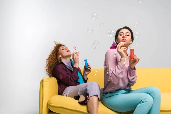 Pelirroja y afroamericana mujer soplando burbujas de jabón mientras se sienta en el sofá amarillo en blanco - foto de stock