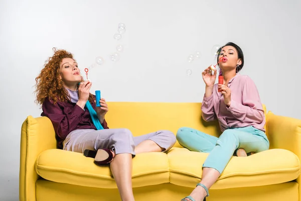 Lockiges rothaariges Mädchen und afrikanisch-amerikanische Frau bläst Seifenblasen, während sie auf einem gelben Sofa auf weiß sitzt — Stockfoto