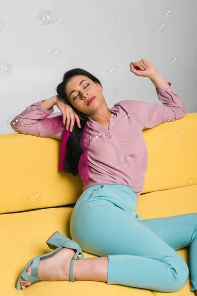 Attraente donna bruna con gli occhi chiusi seduta sul divano vicino a bolle di sapone su bianco — Foto stock