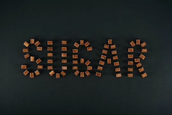 Vue du dessus du mot sucre fait de cubes de sucre brun non raffiné sur fond noir — Photo de stock