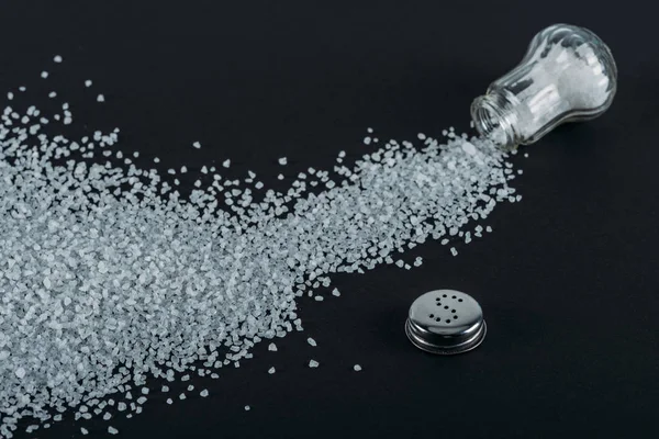 Top view of sprinkled salt crystals near glass salt shaker on black background — Stock Photo