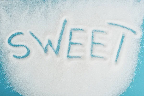 Верхний вид слова сладкий, сделанный на брызганых кристаллах белого сахара на голубой поверхности — стоковое фото