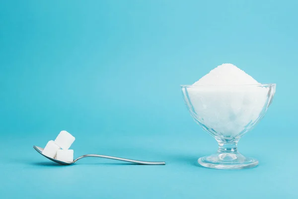 Glass bowl with granulated sugar and teaspoon with sugar cubes on blue background with copy space — Stock Photo